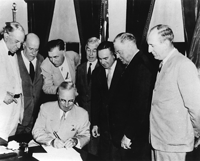 A black and white photo of President Truman sitting with 7 men standing around him while he signs a document.